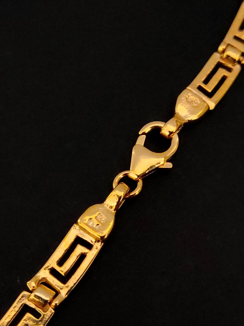 May include: Gold chain necklace with a Greek key pattern and a lobster clasp closure.