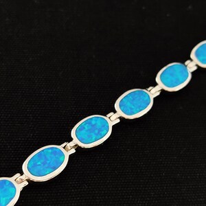 May include: A silver bracelet featuring oval-shaped, vibrant blue opal stones. The opals are set in a silver link chain, creating a delicate and eye-catching piece of jewelry. The bracelet is displayed against a dark background.