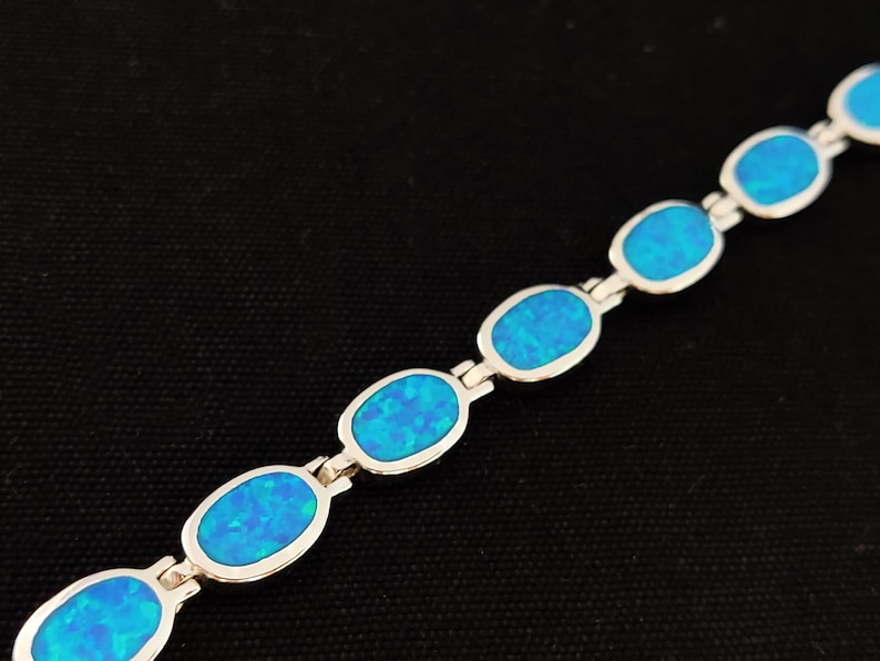 May include: A silver bracelet featuring oval-shaped, vibrant blue opal stones. The opals are set in a silver link chain, creating a delicate and eye-catching piece of jewelry. The bracelet is displayed against a dark background.