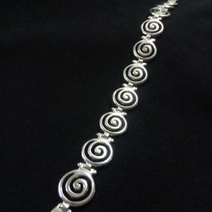 May include: A silver bracelet featuring a series of spiral designs. The bracelet is made of a shiny, metallic material and has a clasp closure. The spirals are evenly spaced along the length of the bracelet, creating a repeating pattern.