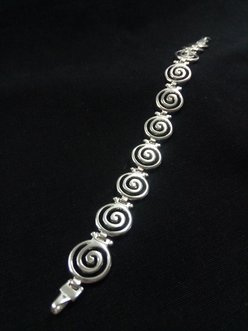 May include: A silver bracelet featuring a series of spiral designs. The bracelet is made of a shiny, metallic material and has a clasp closure. The spirals are evenly spaced along the length of the bracelet, creating a repeating pattern.