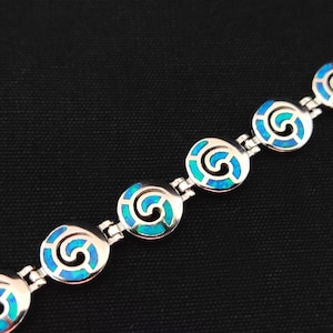 May include: A silver bracelet with blue opal inlay in a spiral pattern. The bracelet is made up of round links with a silver chain connecting them.