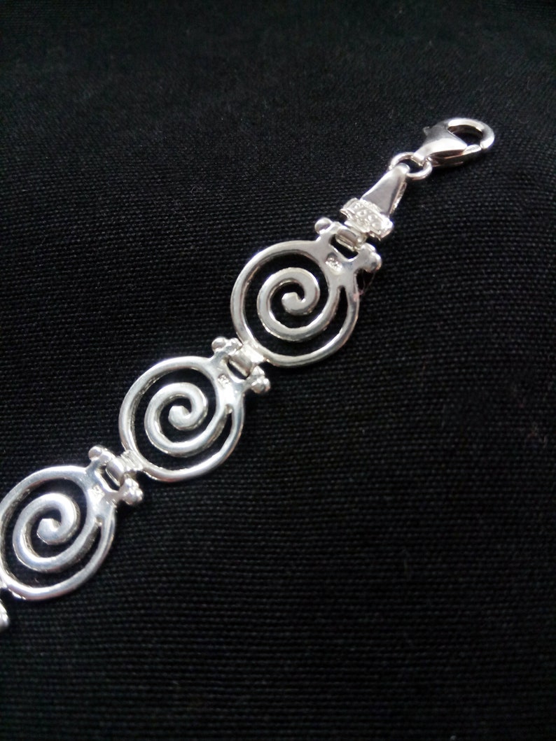 May include: A silver bracelet with a spiral design. The bracelet features three openwork spiral links connected by smaller decorative elements. The clasp is a lobster claw style. The bracelet is displayed against a black background.