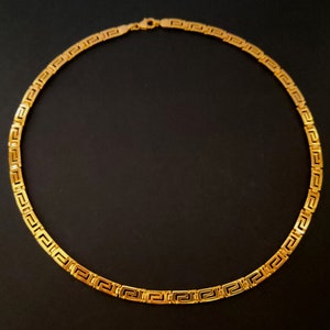 May include: A gold chain necklace with a Greek key pattern. The chain is made of solid gold and has a polished finish.