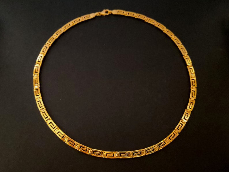 May include: A gold chain necklace with a Greek key pattern. The chain is made of solid gold and has a polished finish.