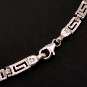 May include: Silver chain necklace with a Greek key pattern and a lobster clasp closure. The chain is marked with the number "925" indicating sterling silver.