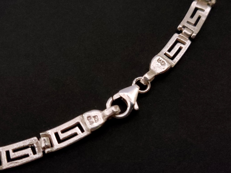 May include: Silver chain necklace with a Greek key pattern and a lobster clasp closure. The chain is marked with the number "925" indicating sterling silver.