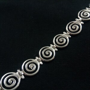 May include: Silver-toned bracelet featuring a repeating spiral design. The bracelet's links are connected by small, decorative bars. The spirals are open and detailed, creating a delicate and elegant look. The jewelry is set against a black background.