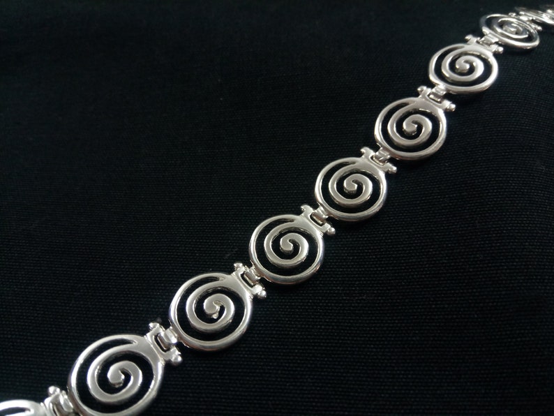 May include: Silver-toned bracelet featuring a repeating spiral design. The bracelet's links are connected by small, decorative bars. The spirals are open and detailed, creating a delicate and elegant look. The jewelry is set against a black background.