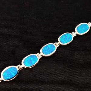 May include: A silver bracelet featuring oval-shaped, blue opal stones. The opals have a vibrant, iridescent quality, set in a linked design. The bracelet is displayed against a black background.