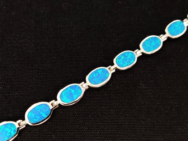May include: A silver bracelet featuring oval-shaped, blue opal stones. The opals have a vibrant, iridescent quality, set in a linked design. The bracelet is displayed against a black background.