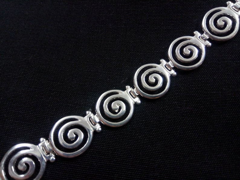 May include: Silver-toned bracelet featuring a series of spiral designs. Each spiral is connected by a small, decorative link. The bracelet is set against a black background, highlighting the bright metal.