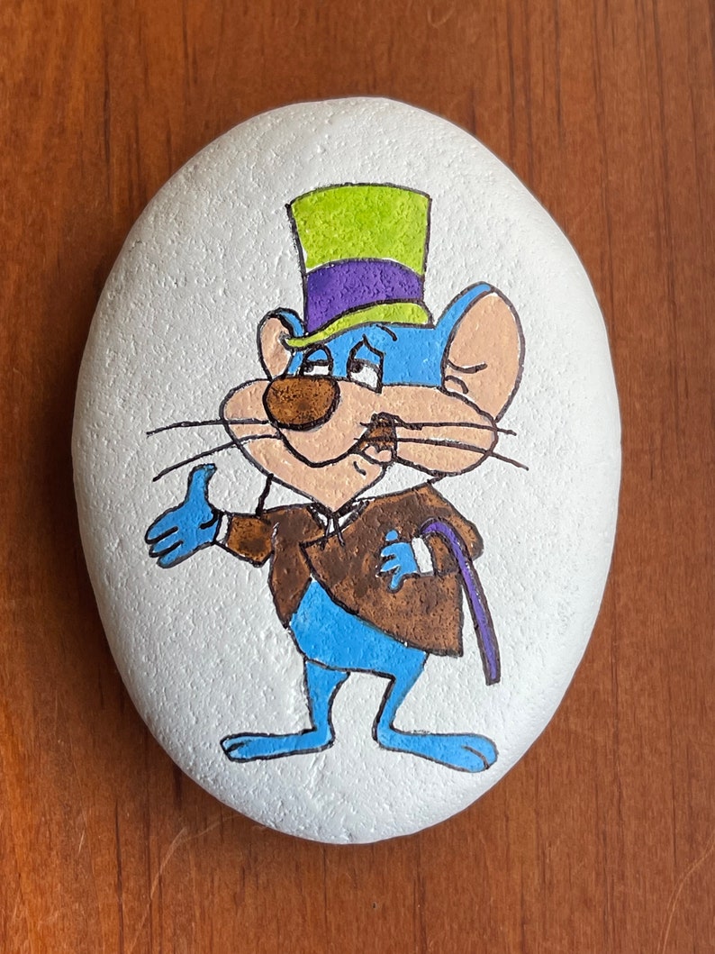 Puede incluir: Piedra pintada a mano con un rat&oacute;n de dibujos animados que lleva un sombrero de copa verde, morado y amarillo. El rat&oacute;n es azul con detalles marrones y sostiene un bast&oacute;n morado.