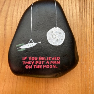 May include: A black painted rock with a white moon and a rocket ship hanging from strings. The rock has the text "If you believed they put a man on the moon." written in pink.