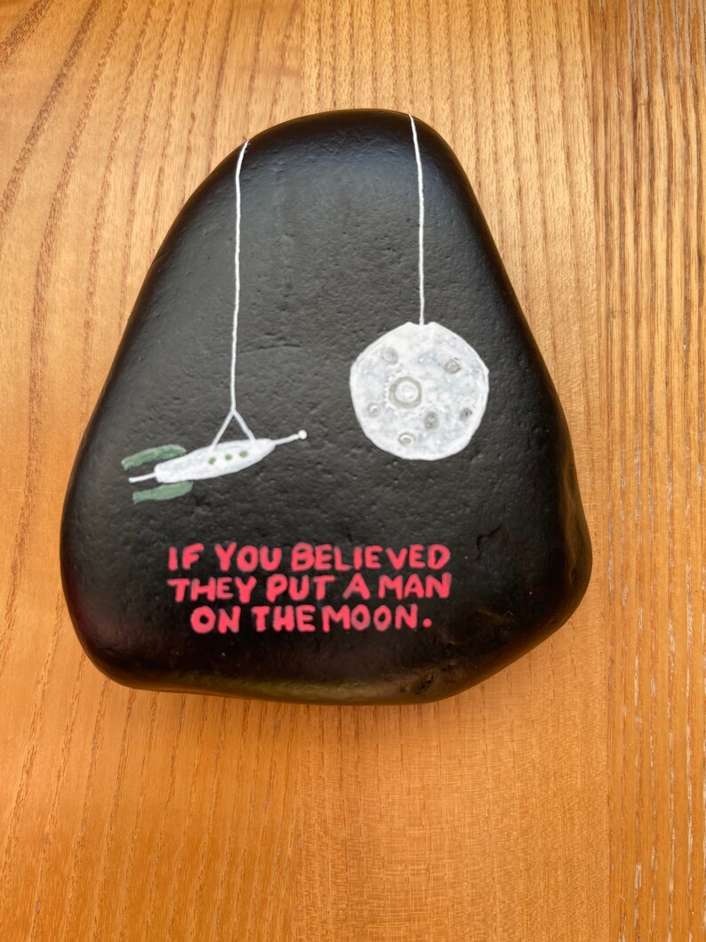 May include: A black painted rock with a white moon and a rocket ship hanging from strings. The rock has the text "If you believed they put a man on the moon." written in pink.
