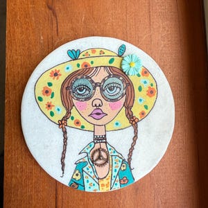 Puede incluir: Una obra de arte redonda pintada a mano que presenta una ilustraci&oacute;n caprichosa de una mujer con coletas, que lleva un sombrero amarillo adornado con flores naranjas y una flor turquesa. Tiene gafas redondas y un collar con el s&iacute;mbolo de la paz. La obra est&aacute; sobre una superficie blanca.