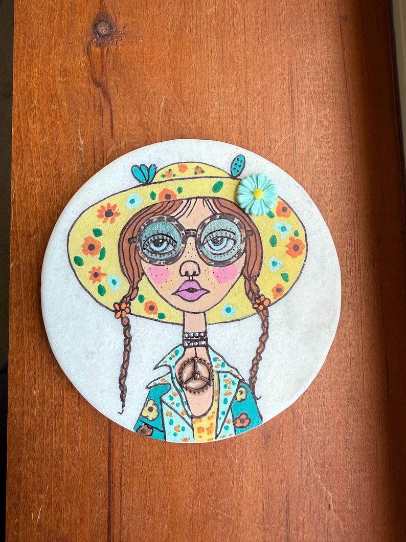 Puede incluir: Una obra de arte redonda pintada a mano que presenta una ilustraci&oacute;n caprichosa de una mujer con coletas, que lleva un sombrero amarillo adornado con flores naranjas y una flor turquesa. Tiene gafas redondas y un collar con el s&iacute;mbolo de la paz. La obra est&aacute; sobre una superficie blanca.