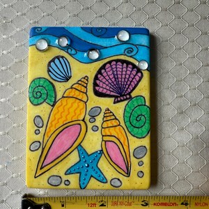 May include: A rectangular, hand-painted tile featuring a beach scene. The design includes yellow sand, blue waves, and various seashells in orange, pink, and purple. A blue starfish and clear, round embellishments add detail. The tile measures approximately 4 inches in length.