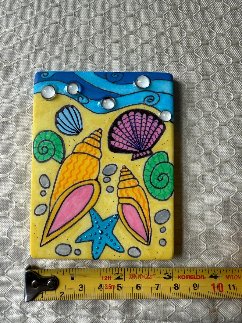 May include: A rectangular, hand-painted tile featuring a beach scene. The design includes yellow sand, blue waves, and various seashells in orange, pink, and purple. A blue starfish and clear, round embellishments add detail. The tile measures approximately 4 inches in length.