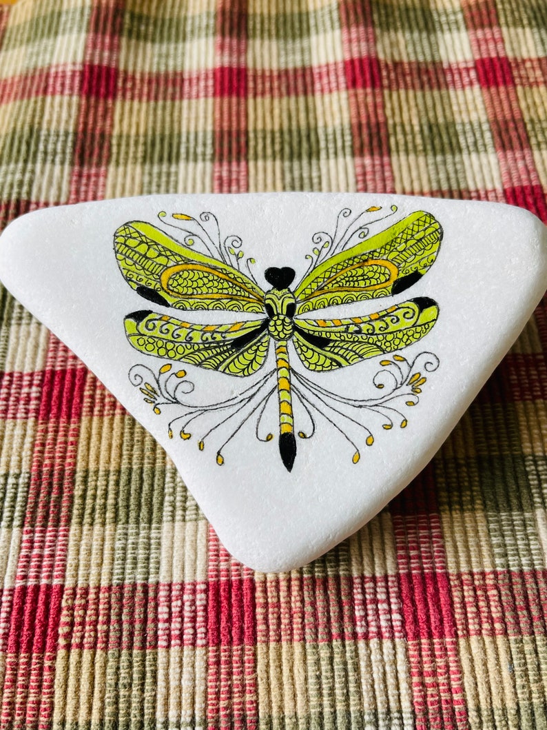 K&ouml;nnte beinhalten: Ein wei&szlig; bemalter Stein mit einem gelben und schwarzen Libellen-Design. Die Libelle hat komplizierte Details und ist im Doodle-Stil bemalt.