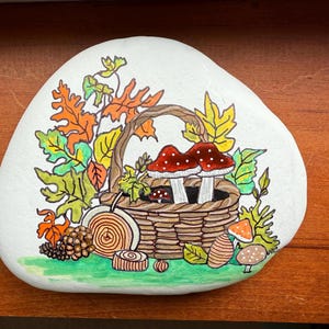 Puede incluir: Piedra blanca pintada a mano con tem&aacute;tica oto&ntilde;al. Una cesta tejida contiene dos setas de sombrero rojo con puntos blancos. Hojas en tonos naranja, amarillo y verde rodean la cesta. Una pi&ntilde;a y otros elementos forestales completan el dise&ntilde;o.