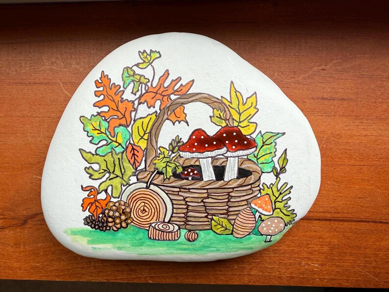 Puede incluir: Piedra blanca pintada a mano con tem&aacute;tica oto&ntilde;al. Una cesta tejida contiene dos setas de sombrero rojo con puntos blancos. Hojas en tonos naranja, amarillo y verde rodean la cesta. Una pi&ntilde;a y otros elementos forestales completan el dise&ntilde;o.