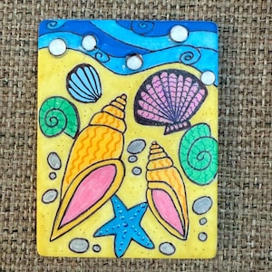 May include: A rectangular painting depicting a beach scene with a blue sky and ocean waves. The artwork features a yellow sandy area with various seashells, a blue starfish, and small gray stones. The shells are in shades of orange, pink, and green.