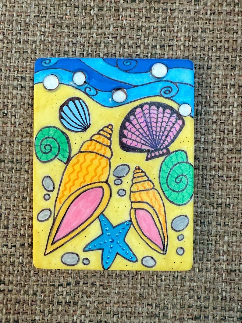 May include: A rectangular painting depicting a beach scene with a blue sky and ocean waves. The artwork features a yellow sandy area with various seashells, a blue starfish, and small gray stones. The shells are in shades of orange, pink, and green.
