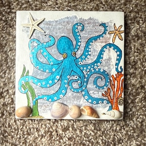 Puede incluir: Obra de arte cuadrada con un pulpo azul con lunares blancos, estrellas de mar, conchas y coral. El pulpo es el punto focal, con sus tentáculos elegantemente curvados. El fondo tiene una apariencia texturizada y descolorida, realzando el tema marino.