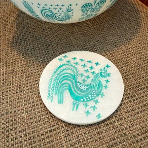 Puede incluir: Un posavasos redondo blanco con un diseño de gallo turquesa y detalles turquesas. El posavasos está colocado sobre una superficie marrón texturizada. Un cuenco blanco con un diseño de gallo turquesa está en el fondo.