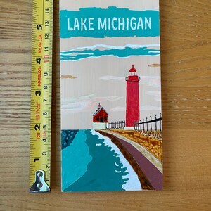 K&ouml;nnte beinhalten: Ein Holzschild mit einer gemalten Szene eines roten Leuchtturms an einem Seeufer. Das Schild tr&auml;gt die Aufschrift "LAKE MICHIGAN" in wei&szlig;en Buchstaben auf einem t&uuml;rkisfarbenen Hintergrund.