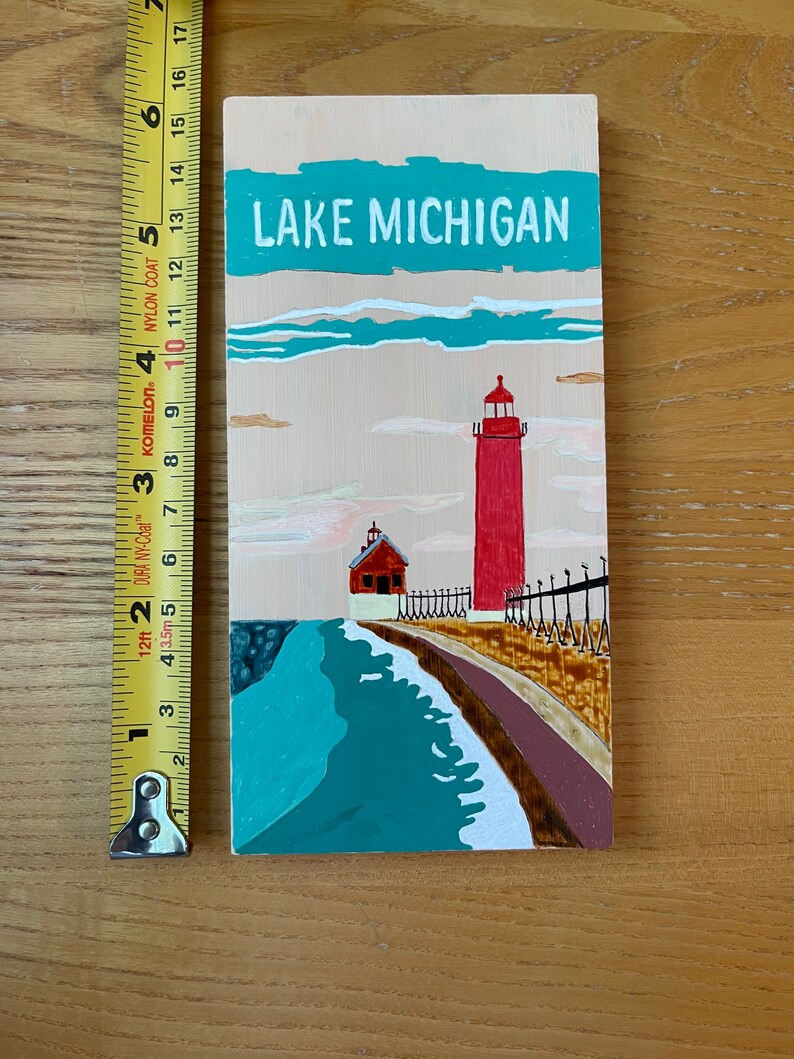 K&ouml;nnte beinhalten: Ein Holzschild mit einer gemalten Szene eines roten Leuchtturms an einem Seeufer. Das Schild tr&auml;gt die Aufschrift "LAKE MICHIGAN" in wei&szlig;en Buchstaben auf einem t&uuml;rkisfarbenen Hintergrund.
