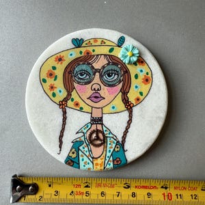 Puede incluir: Un posavasos redondo y blanco con una ilustraci&oacute;n pintada a mano de una mujer con un sombrero floral amarillo y gafas. La mujer tiene trenzas marrones y una chaqueta azul y amarilla. El posavasos mide unos 10 cm de di&aacute;metro.