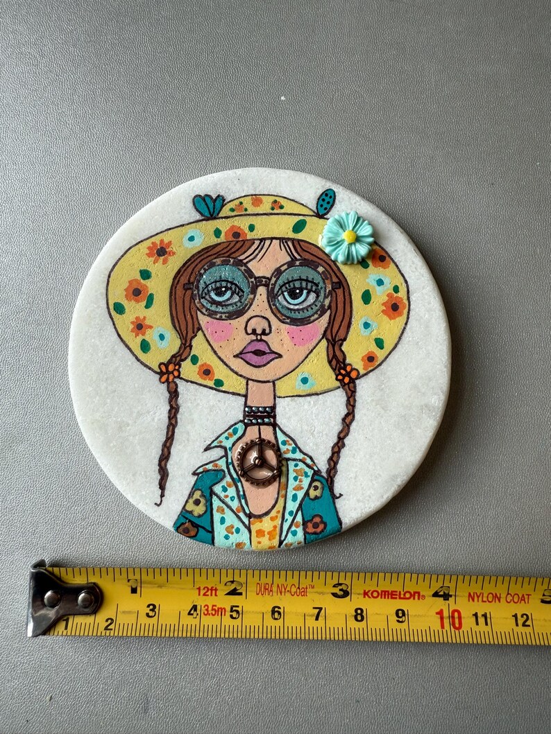 Puede incluir: Un posavasos redondo y blanco con una ilustraci&oacute;n pintada a mano de una mujer con un sombrero floral amarillo y gafas. La mujer tiene trenzas marrones y una chaqueta azul y amarilla. El posavasos mide unos 10 cm de di&aacute;metro.