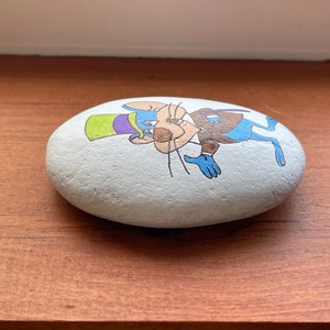 Puede incluir: Una piedra pintada a mano con un rat&oacute;n de dibujos animados que lleva un sombrero verde y morado y un traje azul. El rat&oacute;n est&aacute; pintado en un estilo caprichoso con pelaje marr&oacute;n, vientre blanco y nariz azul.