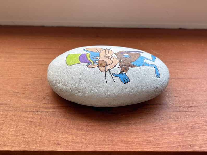 Puede incluir: Una piedra pintada a mano con un rat&oacute;n de dibujos animados que lleva un sombrero verde y morado y un traje azul. El rat&oacute;n est&aacute; pintado en un estilo caprichoso con pelaje marr&oacute;n, vientre blanco y nariz azul.
