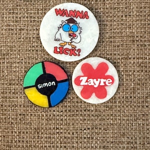 May include: Three colorful round pins. One pin has a cartoon owl with the text "WANNA LICK?". Another pin has the name "Simon" in the center. The third pin has the name "Zayre" in a red flower shape.
