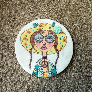Puede incluir: Posavasos redondo con una ilustraci&oacute;n dibujada a mano de una mujer con gafas, un sombrero y un colgante con el s&iacute;mbolo de la paz. El sombrero es amarillo con flores naranjas. La mujer tiene trenzas marrones y una chaqueta turquesa con detalles florales.