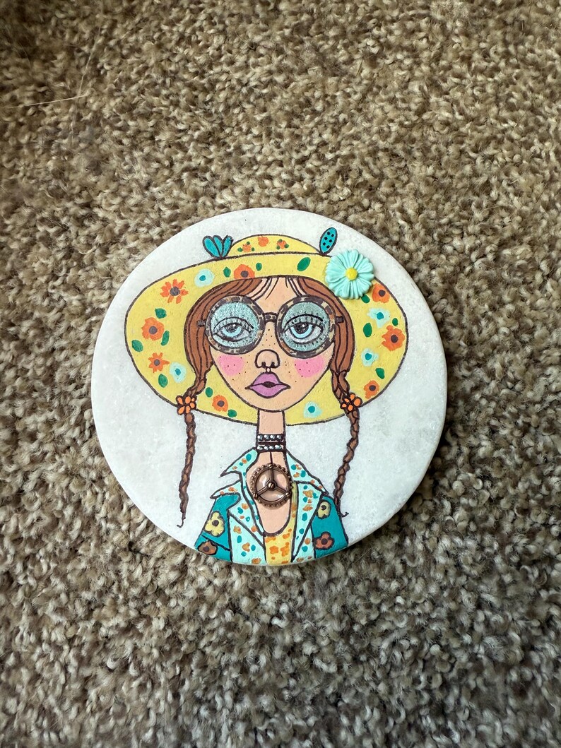 Puede incluir: Posavasos redondo con una ilustraci&oacute;n dibujada a mano de una mujer con gafas, un sombrero y un colgante con el s&iacute;mbolo de la paz. El sombrero es amarillo con flores naranjas. La mujer tiene trenzas marrones y una chaqueta turquesa con detalles florales.