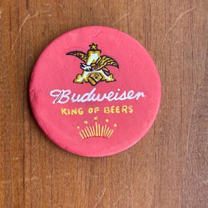 Puede incluir: Un posavasos redondo rojo con un logotipo dorado de Budweiser y el texto "King of Beers".