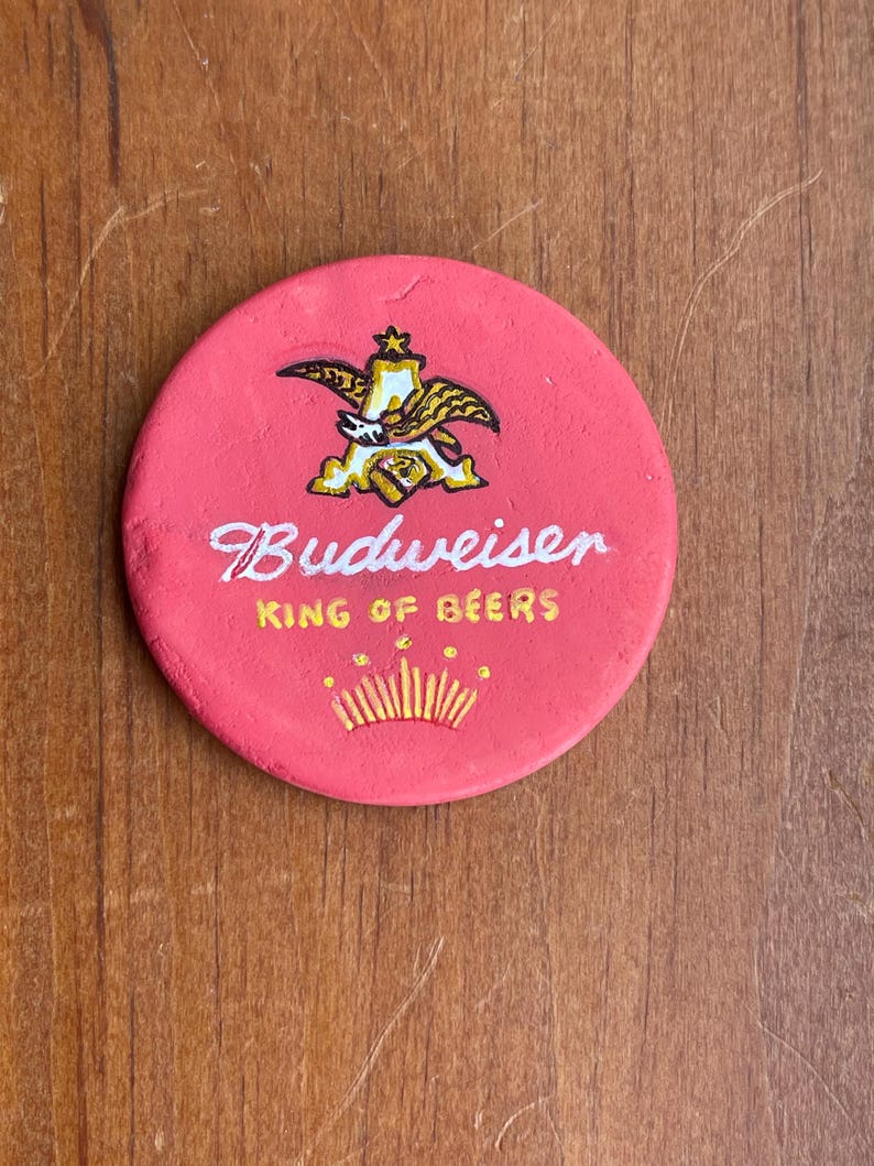 Puede incluir: Un posavasos redondo rojo con un logotipo dorado de Budweiser y el texto "King of Beers".