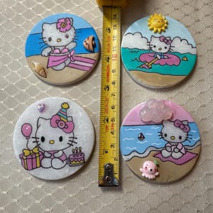 Op de afbeelding: Vier ronde keramische onderzetters met Hello Kitty in verschillende zomerse taferelen. De onderzetters zijn wit met een glinsterende afwerking. De eerste onderzetter toont Hello Kitty in een roze bikini en een bloem in haar haar, staand op een strand met een blauwe zee en een schelp. De tweede onderzetter toont Hello Kitty in een roze reddingsvest, drijvend op een roze zwemband in de zee. De derde onderzetter toont Hello Kitty met een roze feestmuts, die een roze cadeaudoos en een roze taart met kaarsen vasthoudt. De vierde onderzetter toont Hello Kitty zittend op een strandlaken met een roze parasol en een roze octopus.
