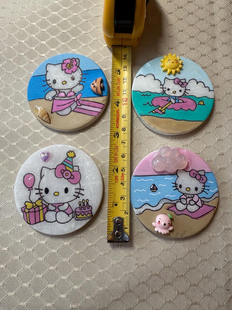 Op de afbeelding: Vier ronde keramische onderzetters met Hello Kitty in verschillende zomerse taferelen. De onderzetters zijn wit met een glinsterende afwerking. De eerste onderzetter toont Hello Kitty in een roze bikini en een bloem in haar haar, staand op een strand met een blauwe zee en een schelp. De tweede onderzetter toont Hello Kitty in een roze reddingsvest, drijvend op een roze zwemband in de zee. De derde onderzetter toont Hello Kitty met een roze feestmuts, die een roze cadeaudoos en een roze taart met kaarsen vasthoudt. De vierde onderzetter toont Hello Kitty zittend op een strandlaken met een roze parasol en een roze octopus.