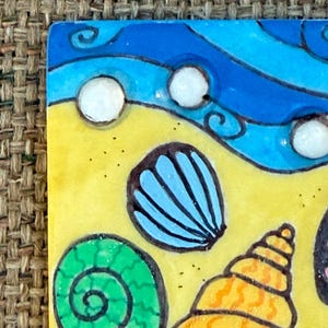 May include: A colorful, square ceramic tile featuring a beach scene. The design includes a yellow sandy area, blue waves, and various seashells in shades of blue, green, and orange. White bubbles are scattered across the blue waves.