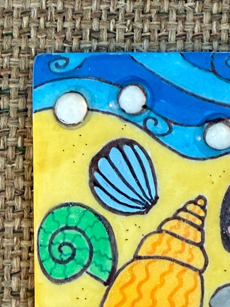 May include: A colorful, square ceramic tile featuring a beach scene. The design includes a yellow sandy area, blue waves, and various seashells in shades of blue, green, and orange. White bubbles are scattered across the blue waves.