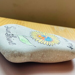 Puede incluir: Una piedra pintada de gris con un girasol amarillo y las palabras "& Sunshine" y "be my" escritas en ella.