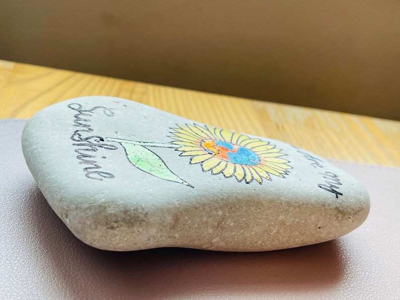 Puede incluir: Una piedra pintada de gris con un girasol amarillo y las palabras "& Sunshine" y "be my" escritas en ella.