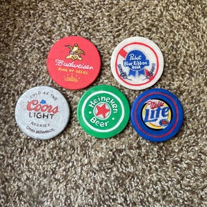 Puede incluir: Cinco tapas redondas de botellas de cerveza de colores con logotipos. Las tapas son rojas, blancas, verdes y azules. Los logotipos incluyen Budweiser, Pabst Blue Ribbon, Coors Light, Heineken y Miller Lite.