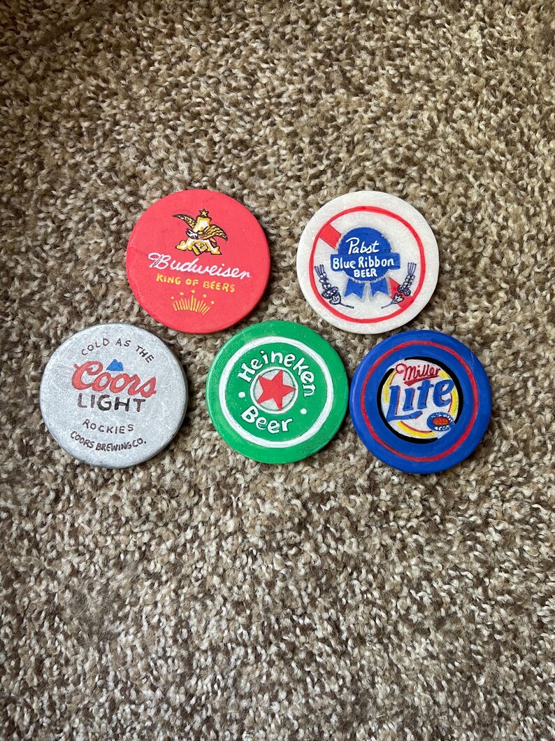 Puede incluir: Cinco tapas redondas de botellas de cerveza de colores con logotipos. Las tapas son rojas, blancas, verdes y azules. Los logotipos incluyen Budweiser, Pabst Blue Ribbon, Coors Light, Heineken y Miller Lite.