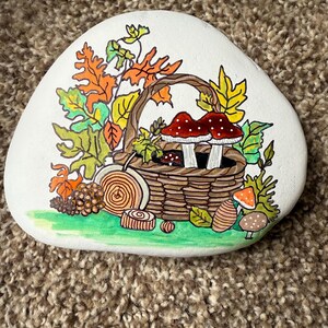 Puede incluir: Una piedra blanca pintada a mano presenta una cesta marr&oacute;n llena de setas con gorro rojo y follaje verde. Hojas de oto&ntilde;o en naranja, amarillo y verde rodean la cesta. Detalles pintados adicionales: caracol, uvas y otras setas.